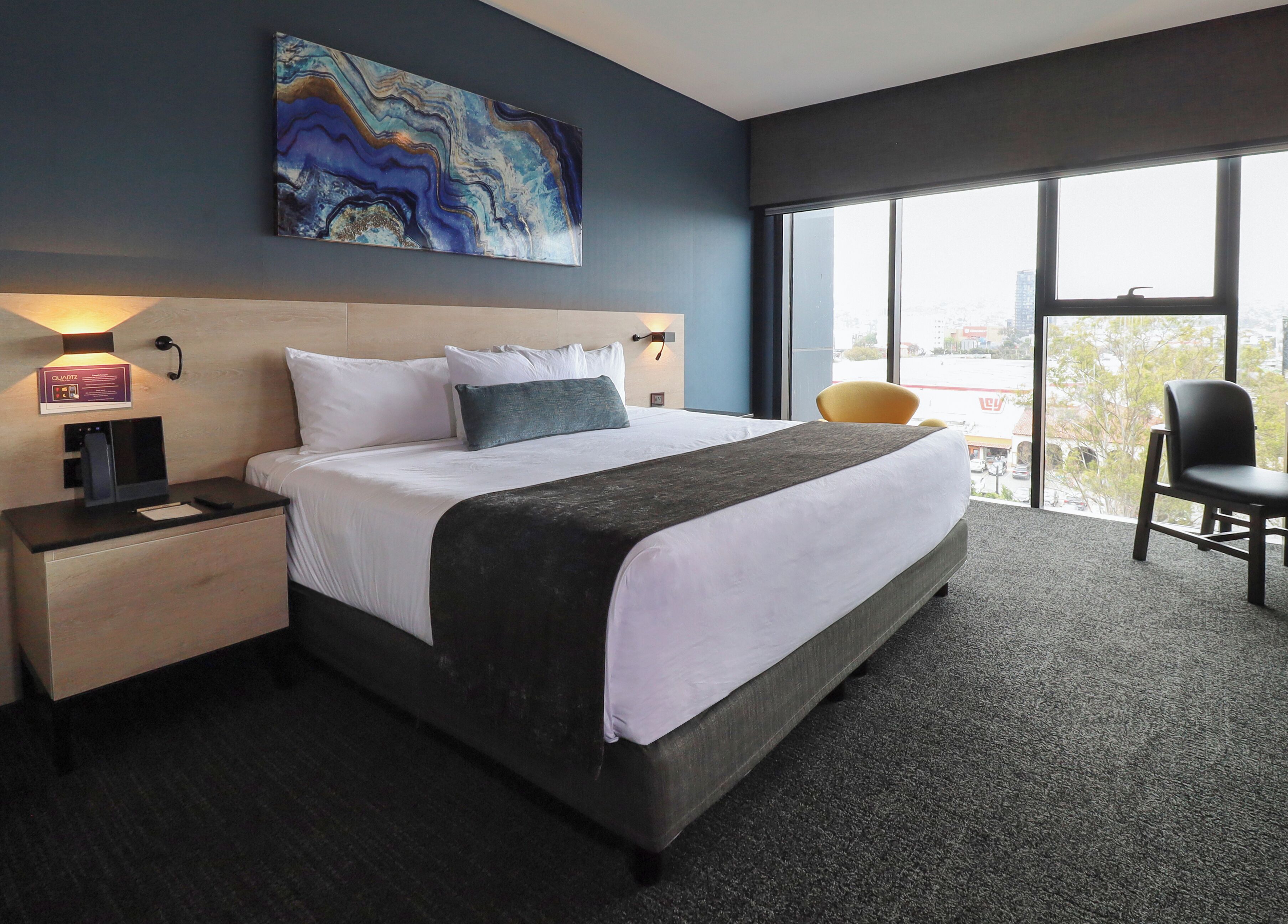 A hotel room with a large bed, a nightstand with a water dispenser, a chair, and a large window offering a city view.