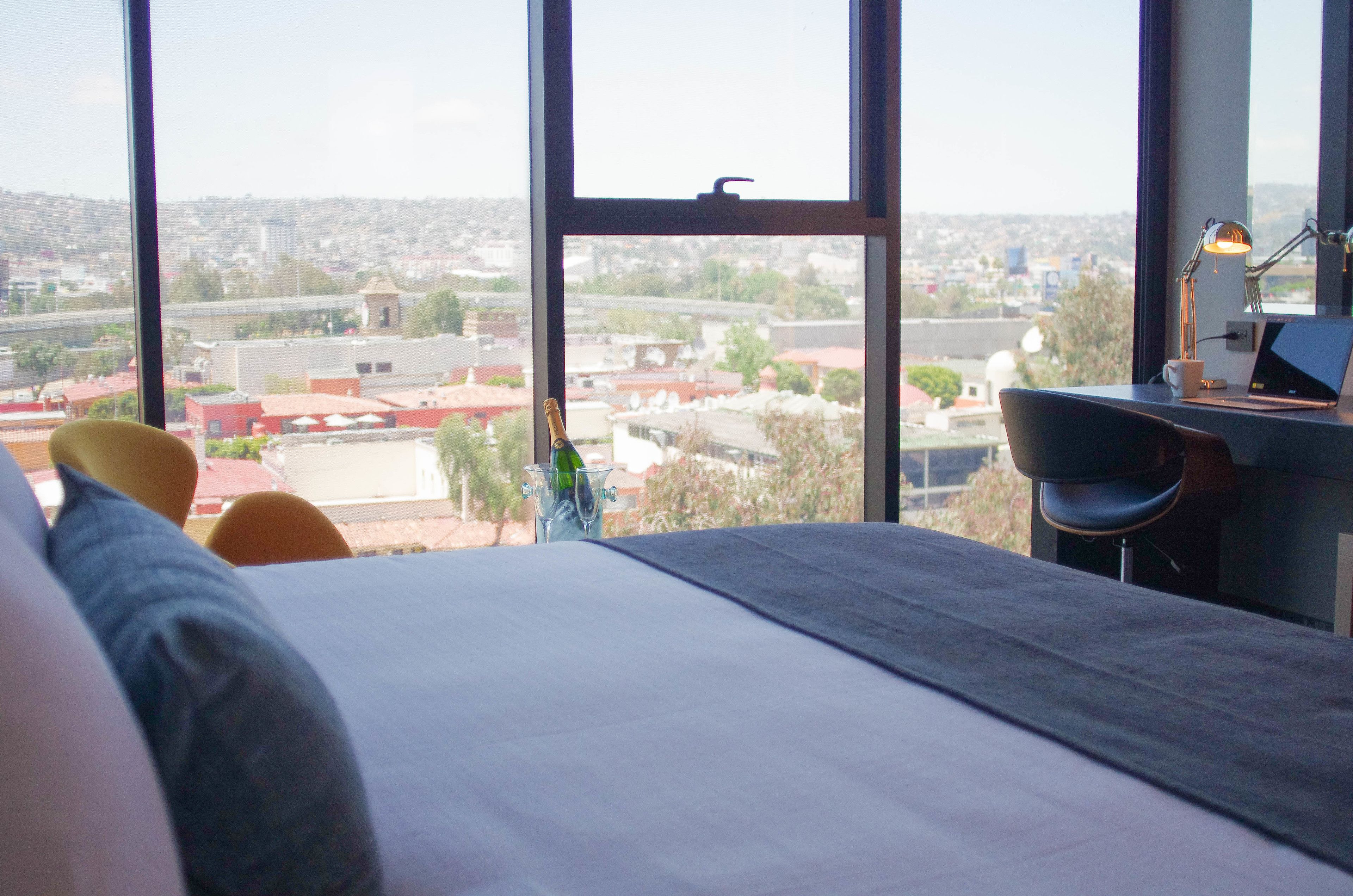 A bedroom with a large window, a bed, a desk with a laptop, and a bottle of champagne.