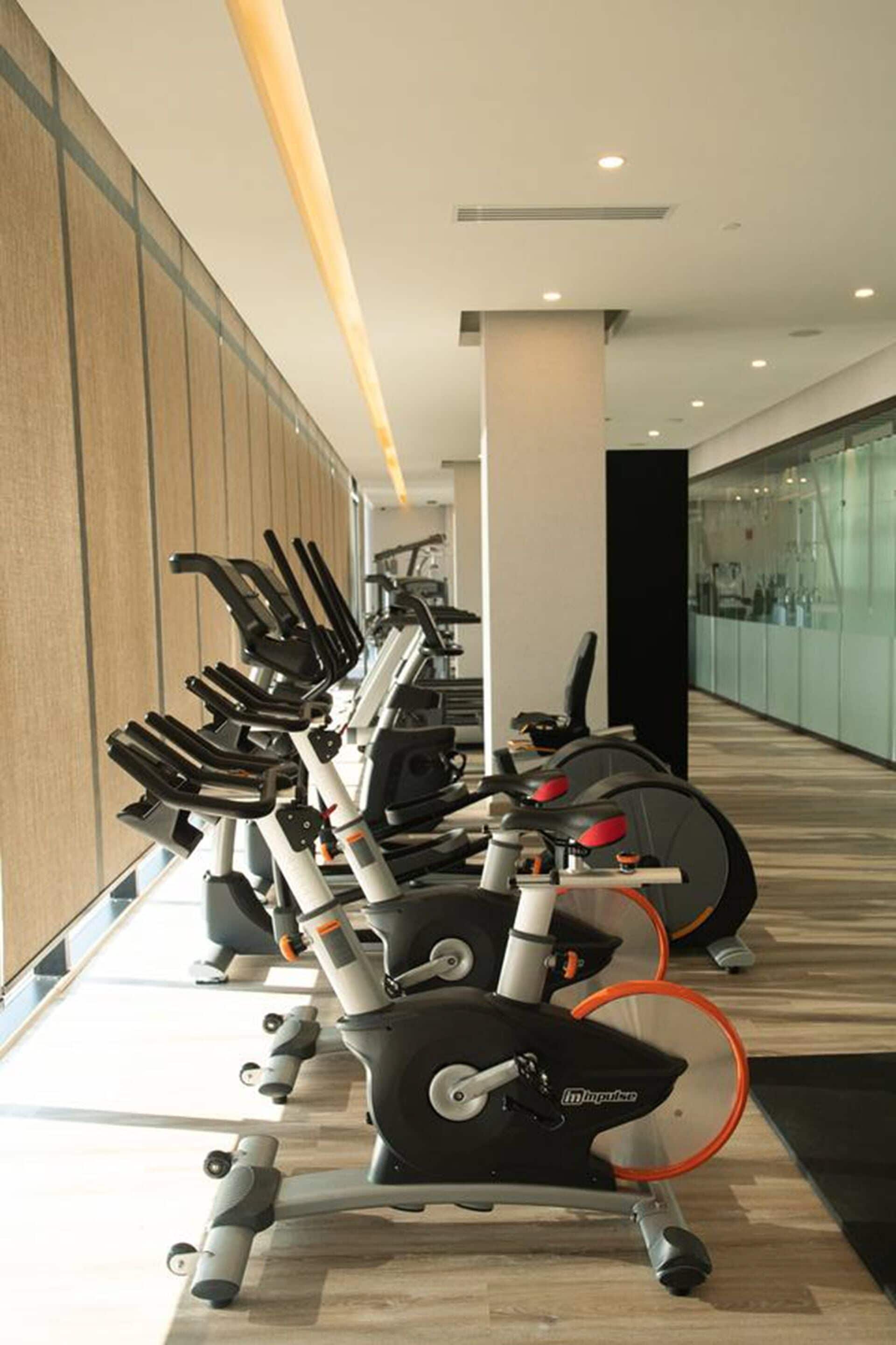A row of stationary bikes in a gym with a wooden floor and glass walls.