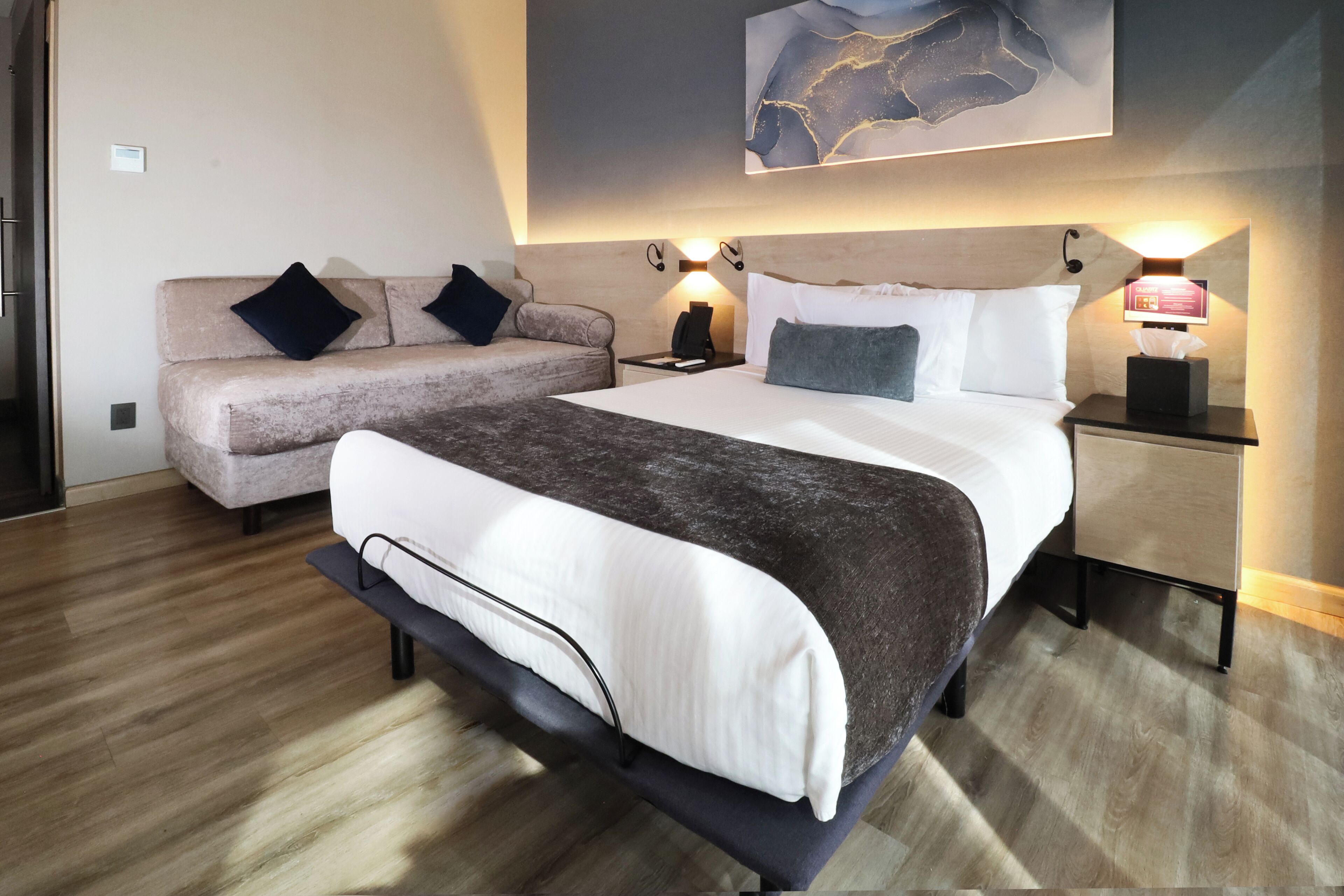 A hotel room with a large bed, a sofa, two bedside tables with lamps, and a wall-mounted artwork.