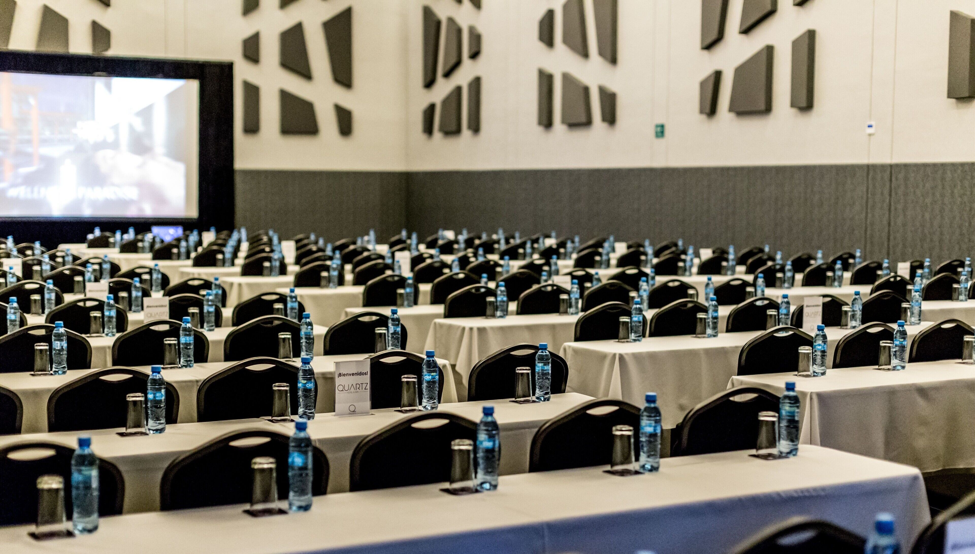 Una sala conferenze con file di tavoli, sedie e bottiglie d'acqua, dotata di cartellini nominativi e uno schermo di proiezione.