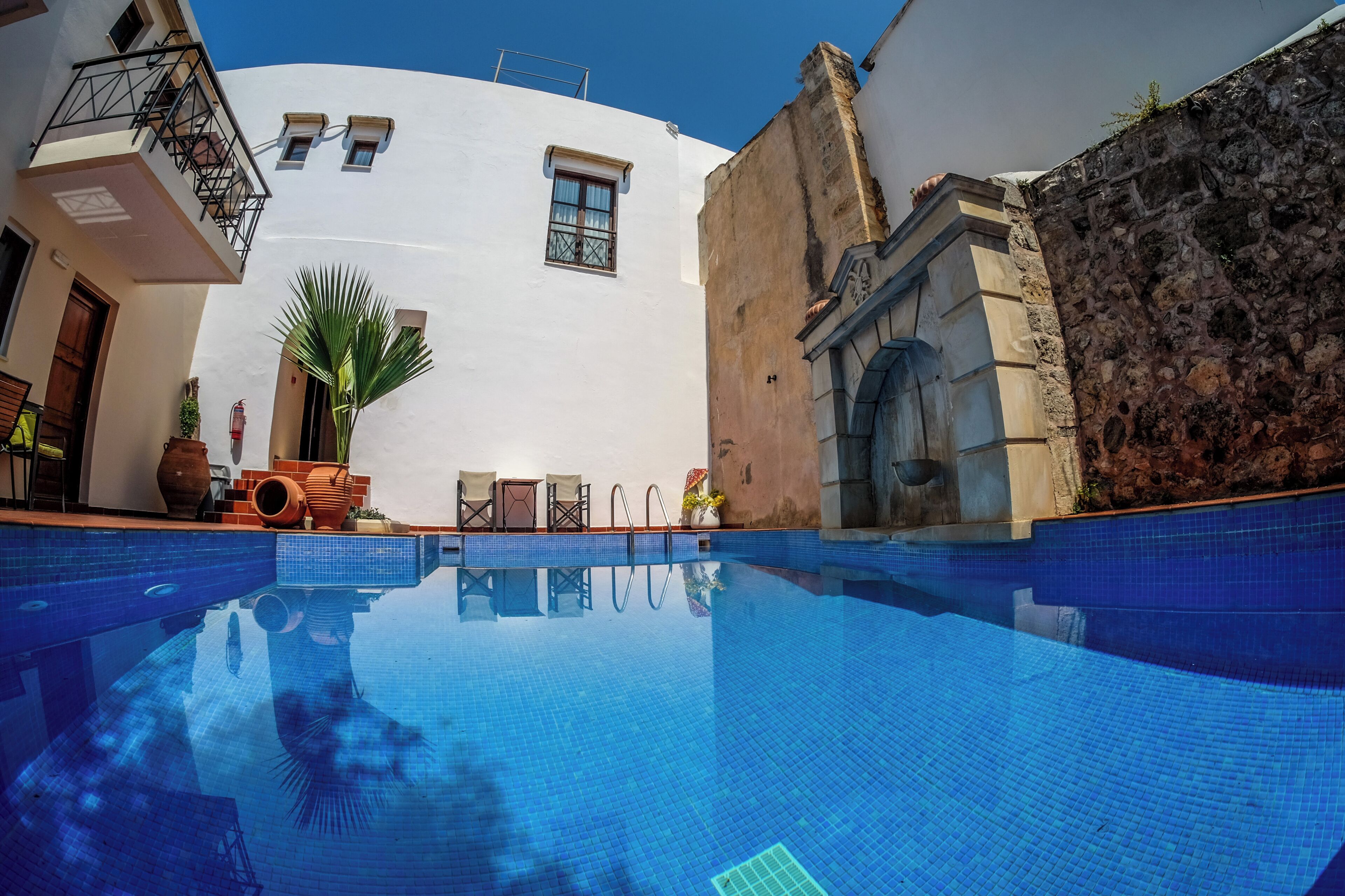A swimming pool with a clear blue water surface, surrounded by a white building with a stone wall, a wooden door, and a small balcony with a metal railing.