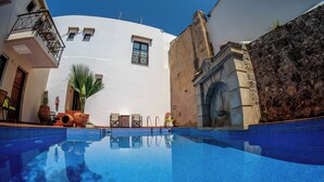 Une piscine à l’eau d’un bleu cristallin, entourée d’un bâtiment blanc doté d’un mur en pierre, d’une porte en bois et d’un petit balcon avec une rambarde en métal.