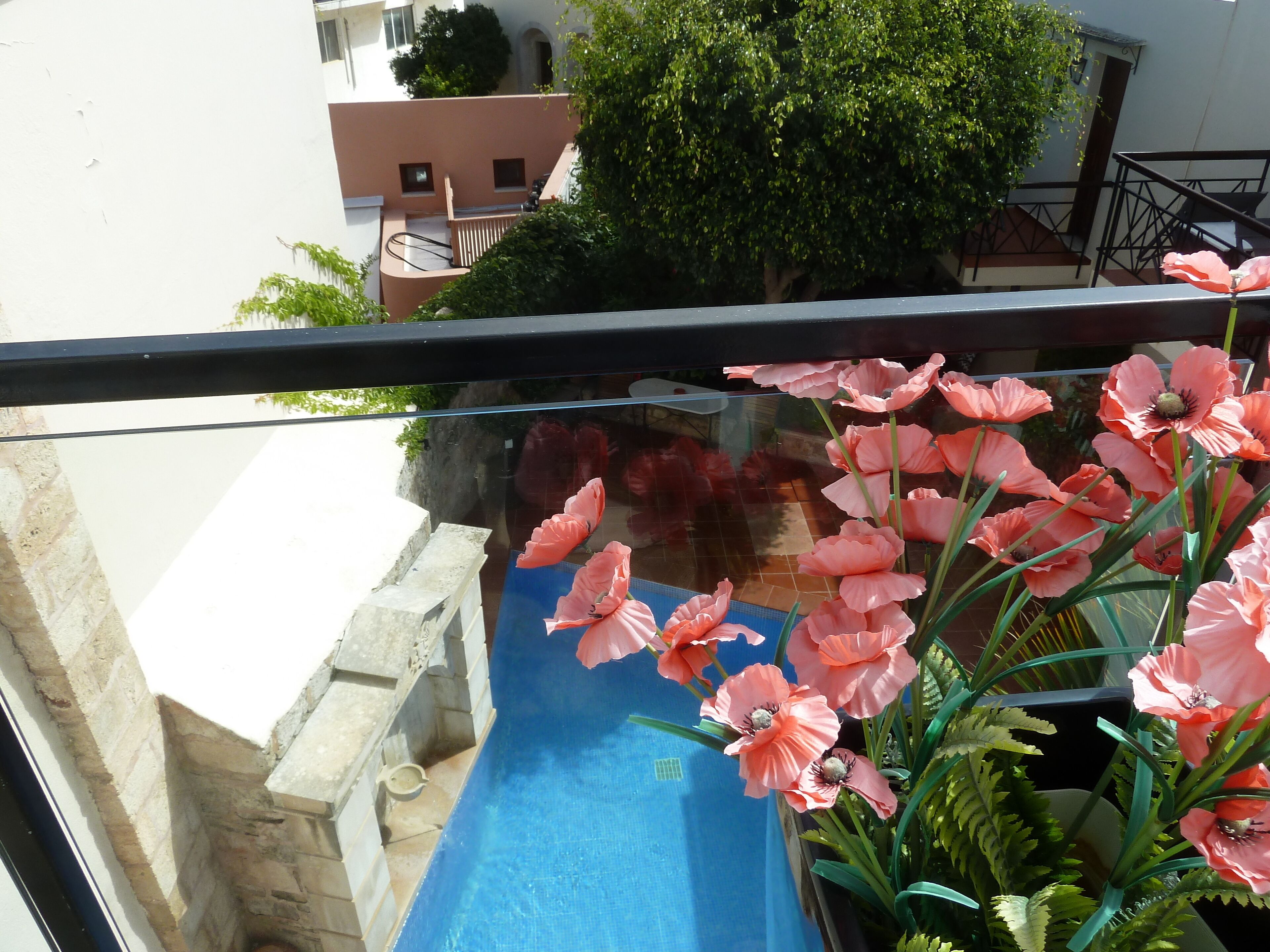 Un balcón con barandilla de vidrio que da a una piscina azul y un arreglo floral rosa.