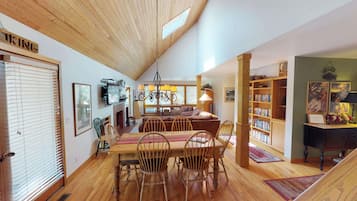 Un salon chaleureux avec un plafond en bois, une cheminée, un coin repas avec des chaises et une table en bois, et une bibliothèque remplie de livres.