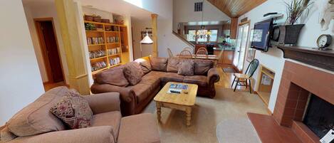 A spacious living area with a large sectional sofa, a coffee table, and a fireplace. There is a kitchen in the background with a dining area.