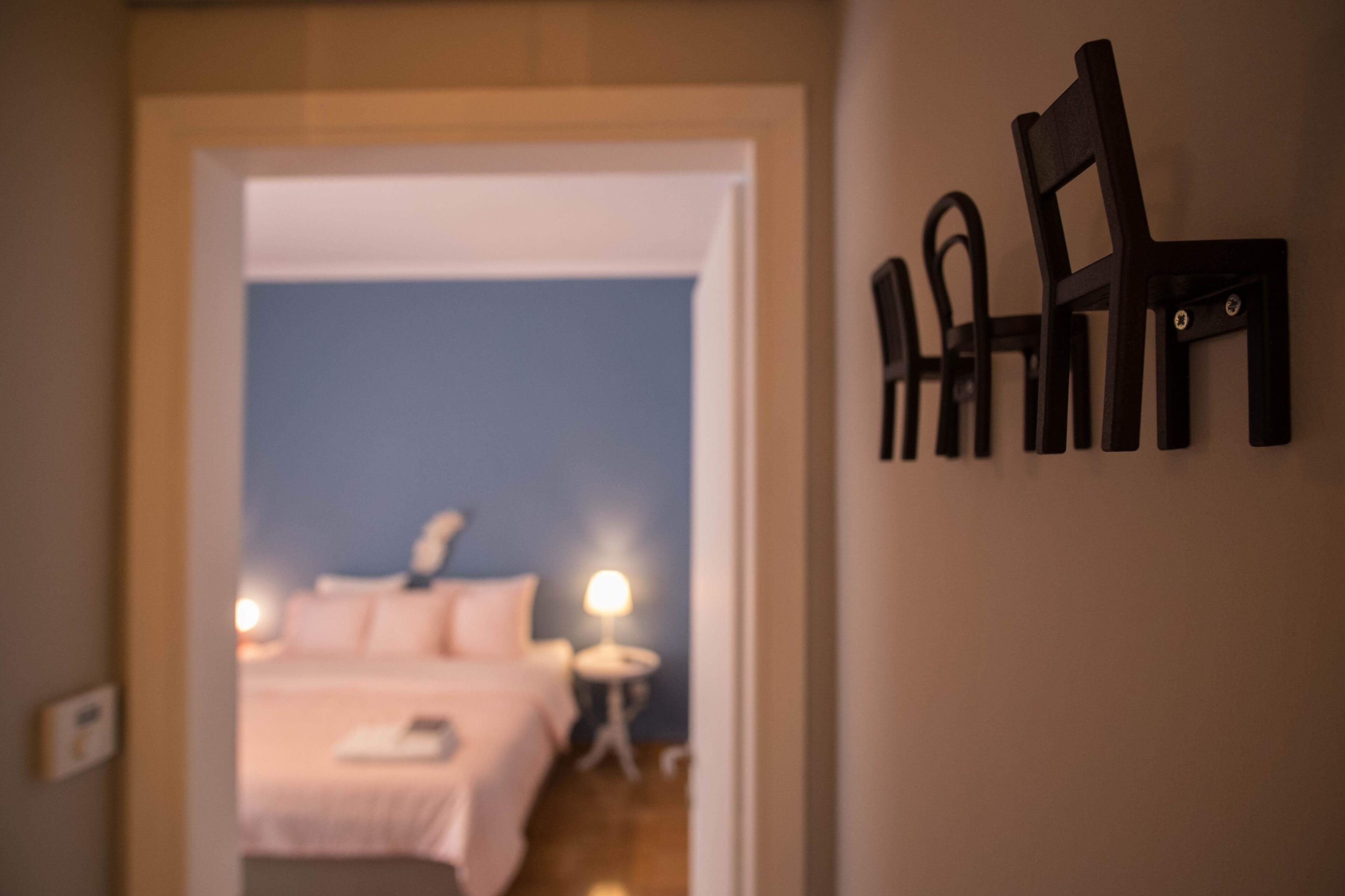 A doorway leading to a bedroom with a bed, a bedside table, and a lamp. There is a wall-mounted chair on the right side of the image.