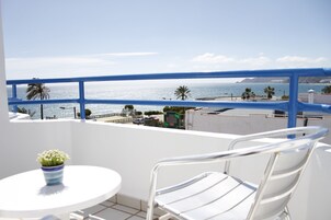 Un balcon avec une table et des chaises blanches, donnant sur une zone côtière bordée de palmiers et offrant une vue sur un ciel dégagé.
