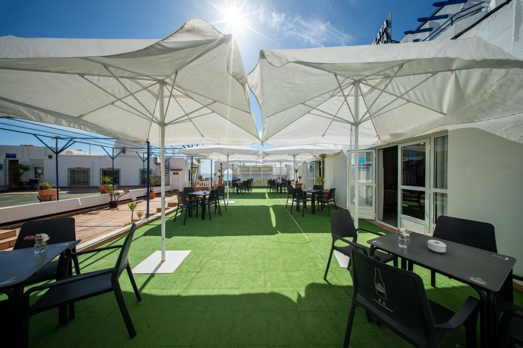 Un patio al aire libre con mesas y sillas, cubierto por grandes sombrillas blancas, y vistas al mar.