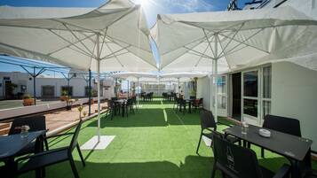 Un patio al aire libre con mesas y sillas, cubierto por grandes sombrillas blancas, y vistas al mar.