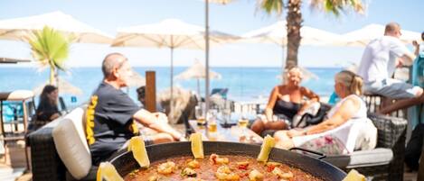 Een eetruimte met een groot visgerecht op de voorgrond, zitplaatsen buiten met parasols, en uitzicht op de zee op de achtergrond.