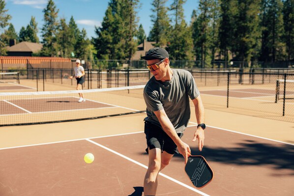 Un court de tennis avec un filet, un joueur portant une casquette et des lunettes de soleil, et un autre joueur à l’arrière-plan.