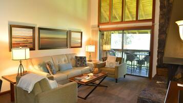 A cozy living room with a sofa, armchairs, a coffee table, and a fireplace. There are large windows and a view of the outdoors.
