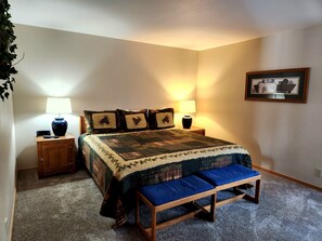 Une chambre avec un grand lit, deux tables de chevet avec des lampes, un banc au pied du lit et un tableau encadré au mur.