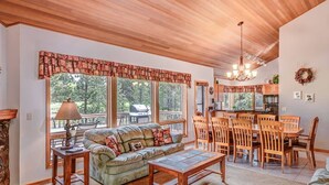 A cozy living room with a sofa, coffee table, and dining area with wooden chairs. Large windows offer a view of the outdoors. - Family Golf Escape with Access to The Cove (Sunriver)