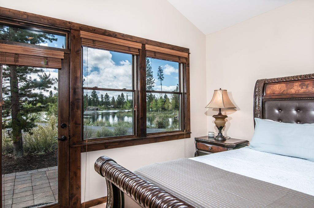 Une chambre avec une grande fenêtre donnant sur un lac et des arbres, un lit avec une tête de lit, une table de chevet avec une lampe et un banc en bois.