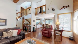A spacious living area with a wooden staircase, a dining area with a table and chairs, and a kitchen visible through an open door. - Cozy Cabin Stay with Access to Exclusive Golf (Sunriver)