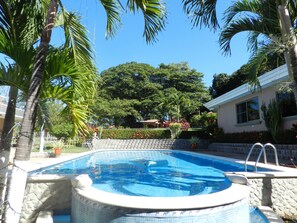 A swimming pool surrounded by palm trees and a stone wall, with a view of a house and lush greenery in the background. - Leave the Cold behind, Winter in the Safe Quiet Paradise of Costa Rica (N/A)