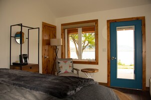 Une chambre à coucher comprenant un lit, une chaise, une petite table, une lampe, un miroir et une porte donnant sur un balcon avec vue sur des arbres et un plan d’eau.