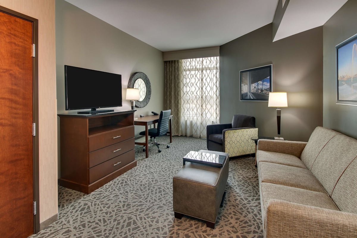 A hotel room with a flat-screen TV, a desk with a chair, a sofa, a patterned carpet, and a large window with curtains.