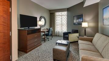 Habitación de hotel con televisión de pantalla plana, un escritorio con una silla, un sofá, alfombra estampada y un ventanal con cortinas.