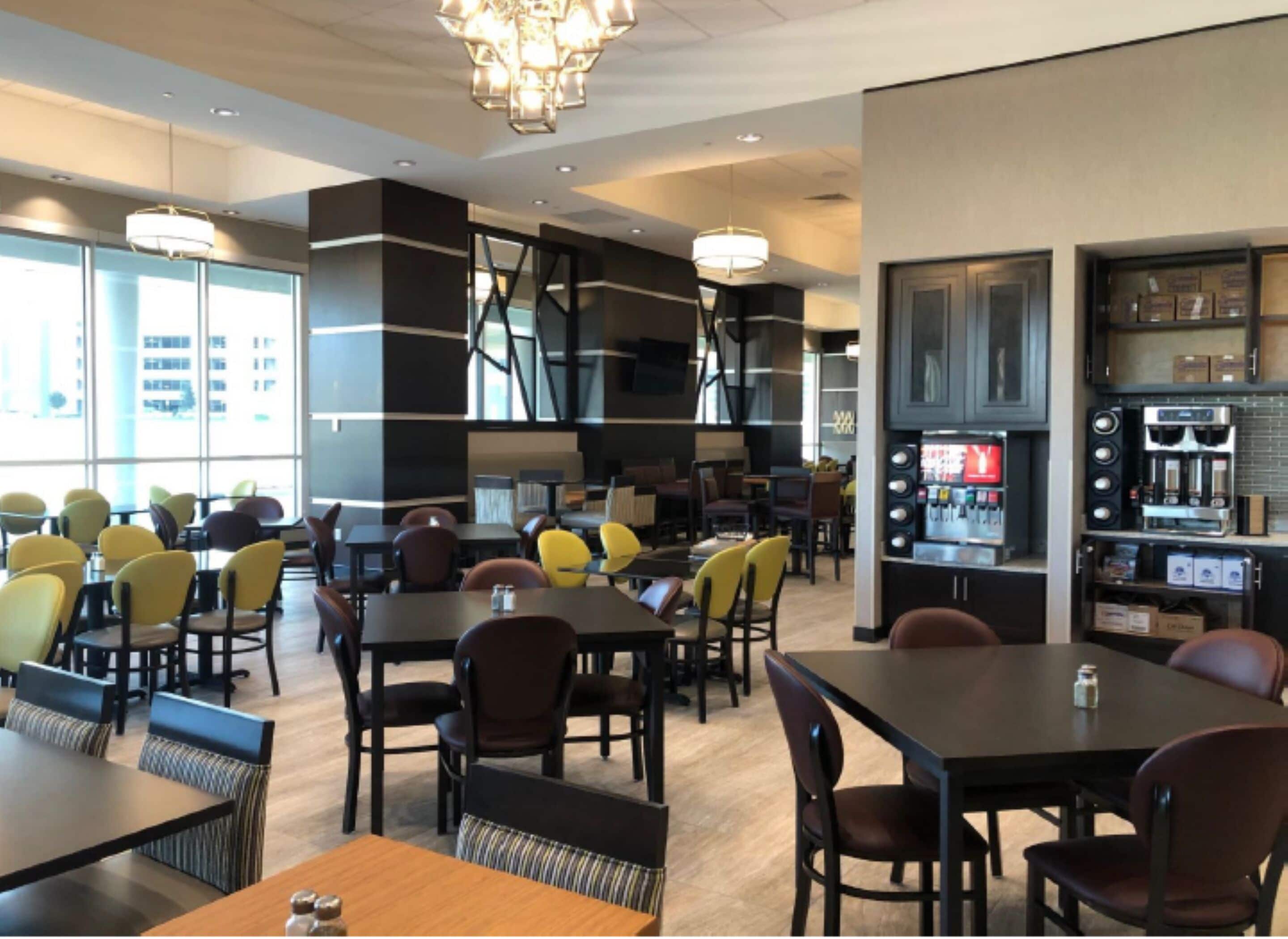 A modern restaurant with multiple dining tables and chairs, a coffee machine, and a large window providing natural light.