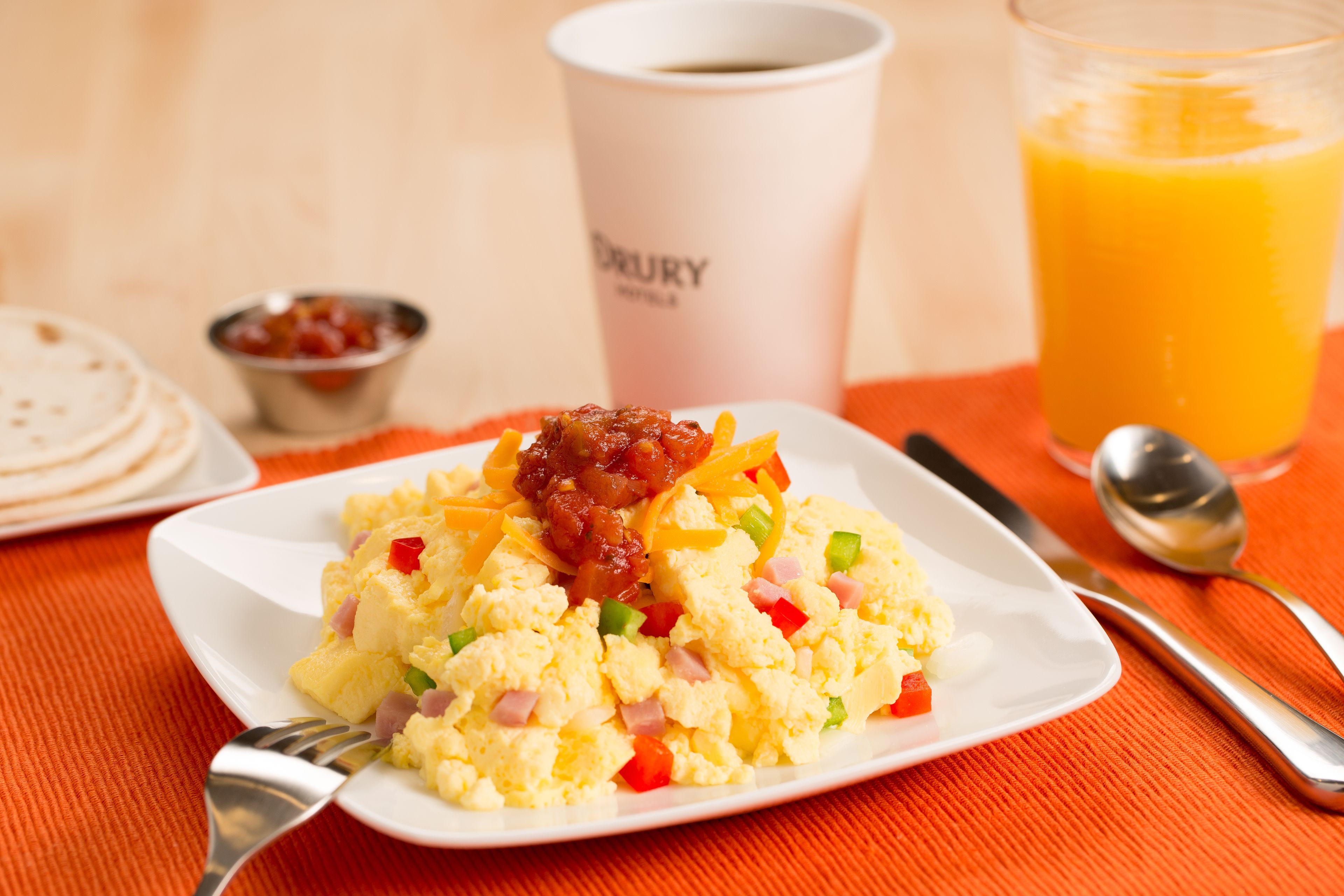Un plato de desayuno con huevos revueltos, jamón y pimientos, una taza de café y un vaso de jugo de naranja sobre un mantel naranja.