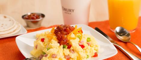 Un plato de desayuno con huevos revueltos, jamón y pimientos, una taza de café y un vaso de jugo de naranja sobre un mantel naranja.