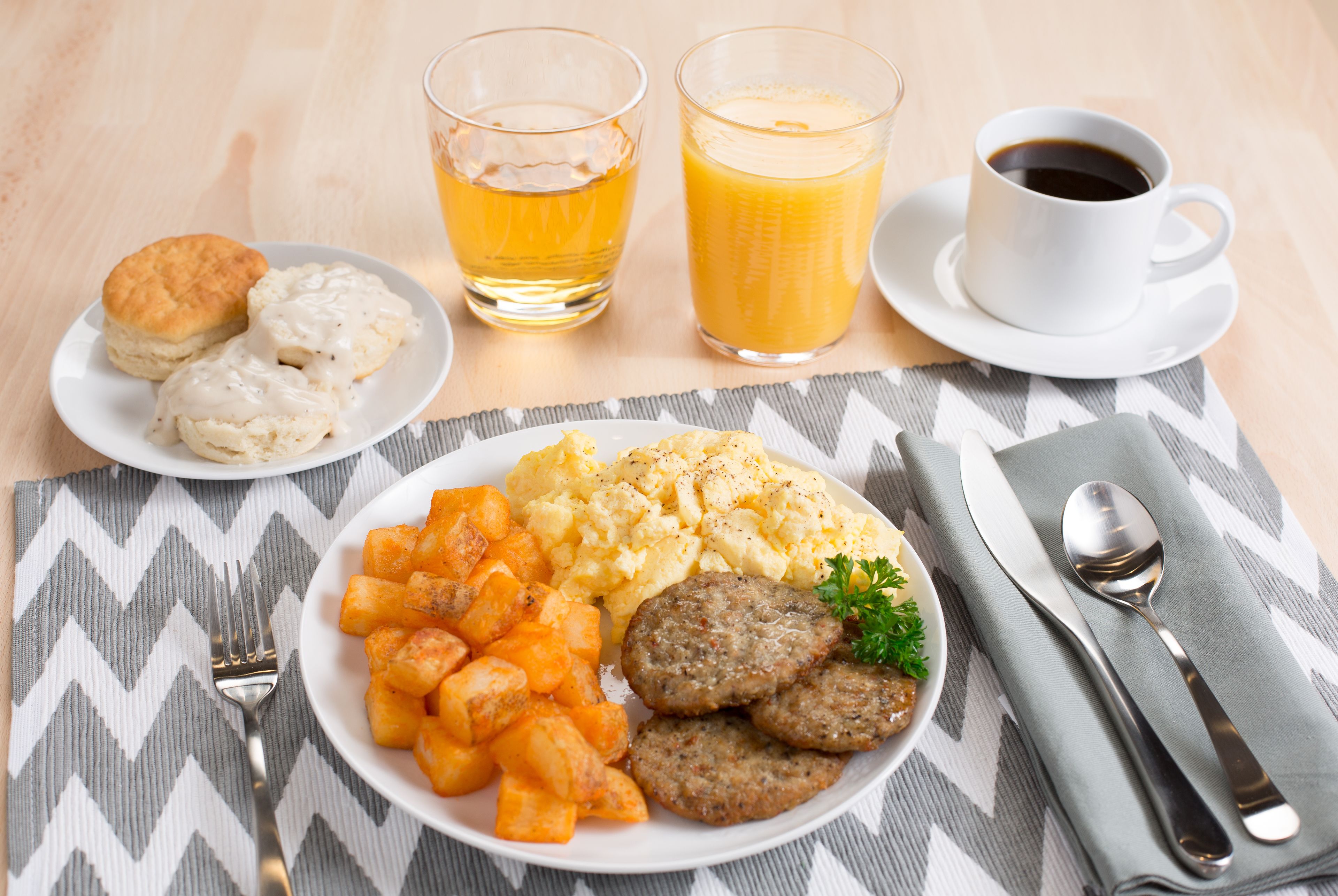 Un plato de desayuno con galletas, huevos revueltos, tortitas de patata y salchichas, acompañado de dos vasos de zumo de naranja y una taza de café.
