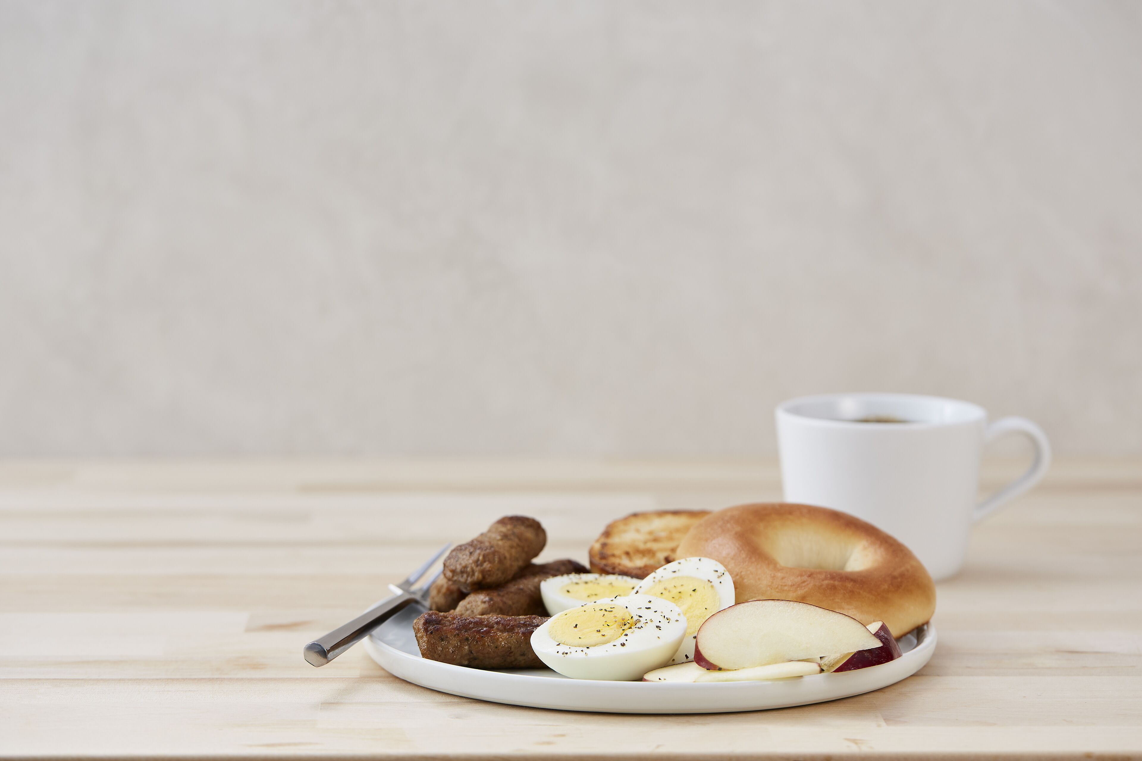 Un plato con un bagel, huevos duros en rodajas, salchichas y rodajas de manzana sobre una mesa de madera.