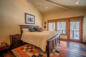 A bedroom with a large bed, a wooden headboard, a quilted rug, a nightstand, a framed picture on the wall, and a window with a view of trees. - Riverside Escape with Access to The Cove (Sunriver)