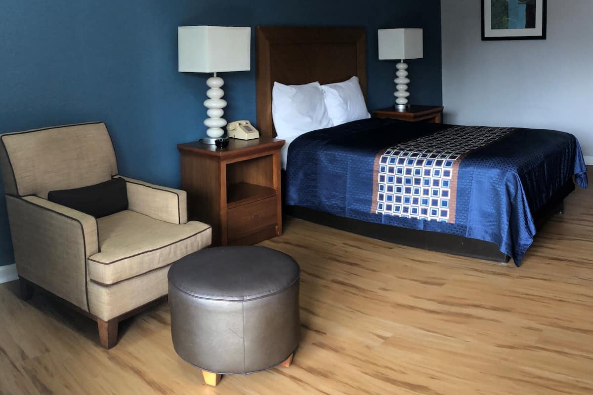 Quarto de hotel com cama, cadeira, mesa de cabeceira, telefone e um pequeno banquinho.