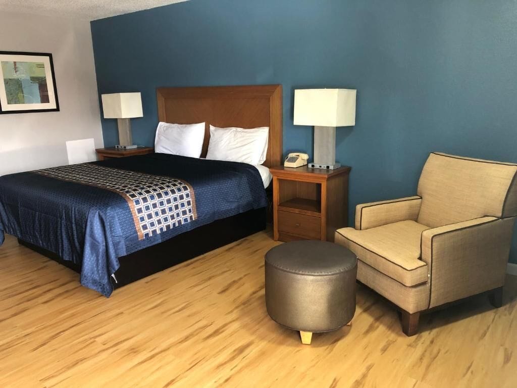 Una camera d'albergo con un letto, due comodini con lampade, una sedia e un poggiapiedi rotondo.