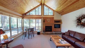 A cozy living room with a fireplace, a television, and a large window overlooking a forest. - Lodge Style Escape with Access to The Cove (Sunriver)
