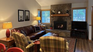 A living room with a fireplace, a sofa, a television, and a window with a view of trees. - Cozy Family Cabin with Access to Exclusive Golf (Sunriver)