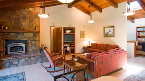 A cozy living room with a stone fireplace, wooden ceiling beams, a red sofa, and a dining table with chairs. - Calm River Oasis with Access to The Cove (Sunriver)