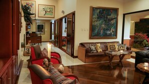 A spacious living room with a brown leather sofa, red armchairs, and a coffee table. There are paintings on the walls and a large window with a view of the outdoors. - Hostal Estancia El Caney (Pereira)