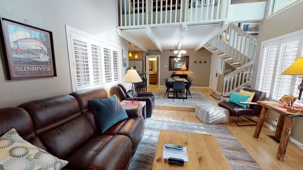 Ein großzügiges Wohnzimmer mit braunem Ledersofa, einem Holztisch und einem Essbereich mit Tisch und Stühlen. Im Hintergrund ist eine Treppe mit Geländer zu sehen.