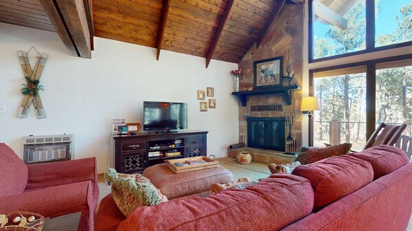 A cozy living room with a stone fireplace, a television, and a large window overlooking trees. - Wrap-Around Deck | Fireplace | Ward Cabin (Angel Fire)