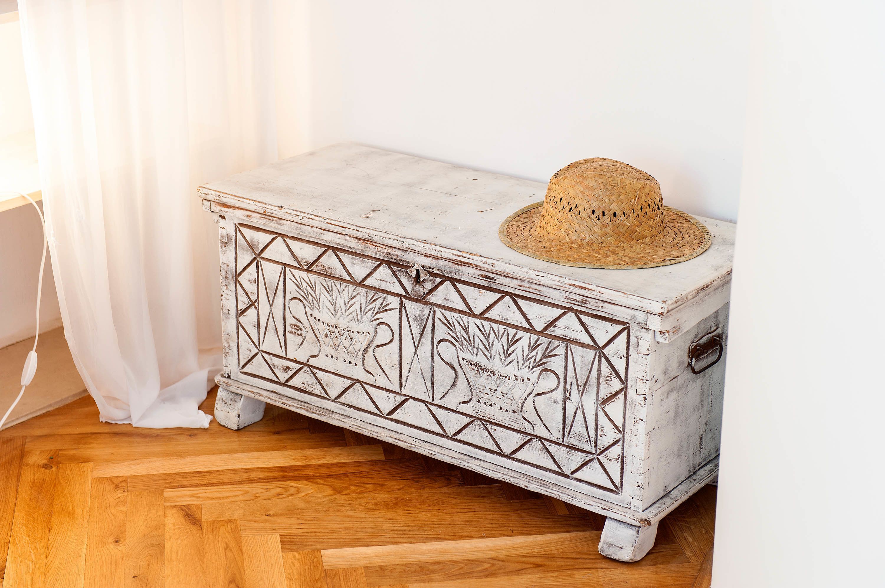 Un cofre de madera blanca con un aspecto envejecido, adornado con patrones geométricos y un sombrero de paja encima.