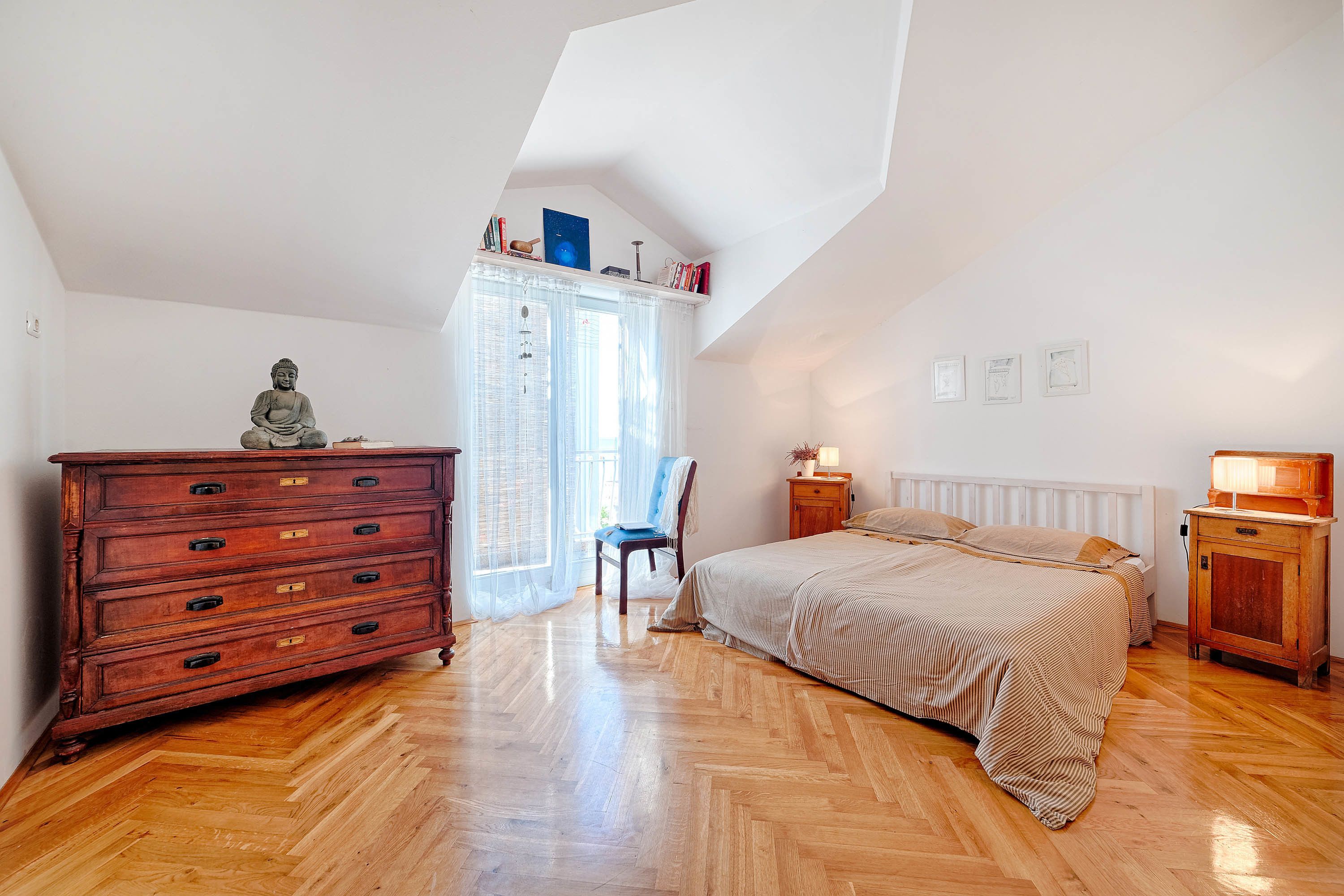 Een slaapkamer met een houten dressoir, een bed met een gestreept dekbedovertrek, een stoel en een nachtkastje met een lamp.
