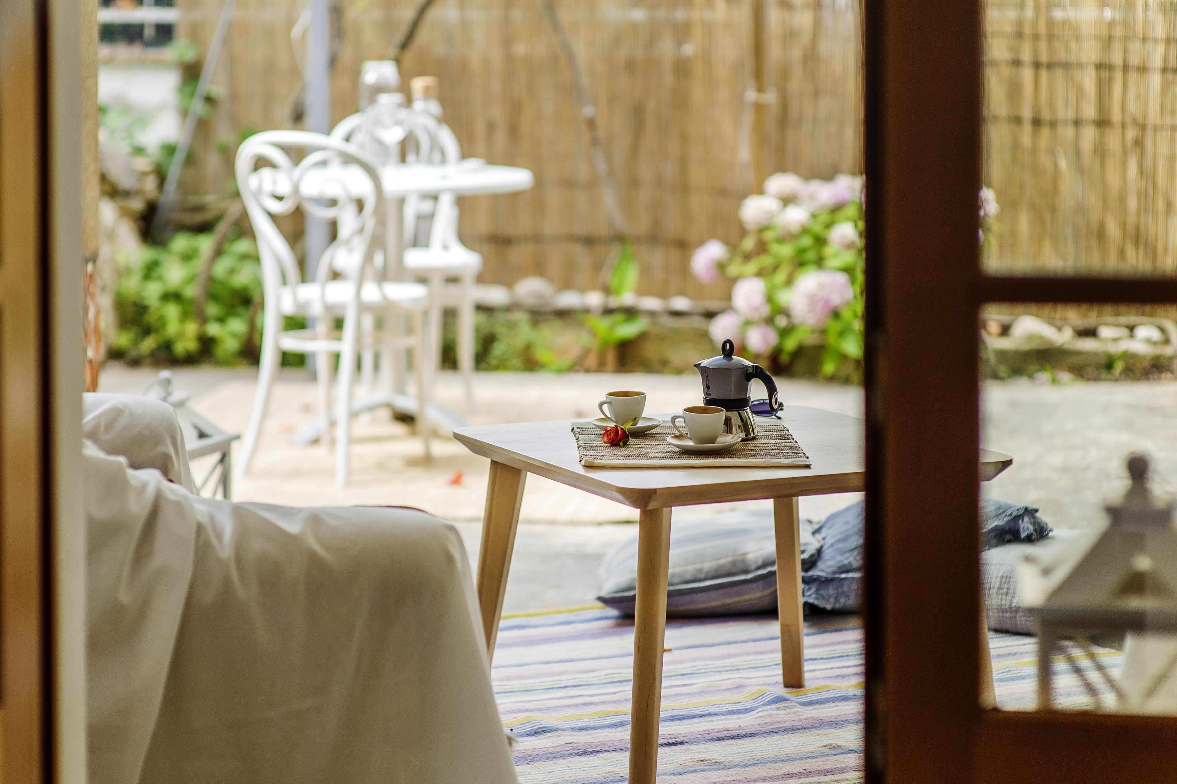 A room with a wooden table set for tea, white chairs, and a view of a garden with pink flowers and a bamboo fence.