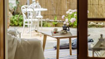 A room with a wooden table set for tea, white chairs, and a view of a garden with pink flowers and a bamboo fence.