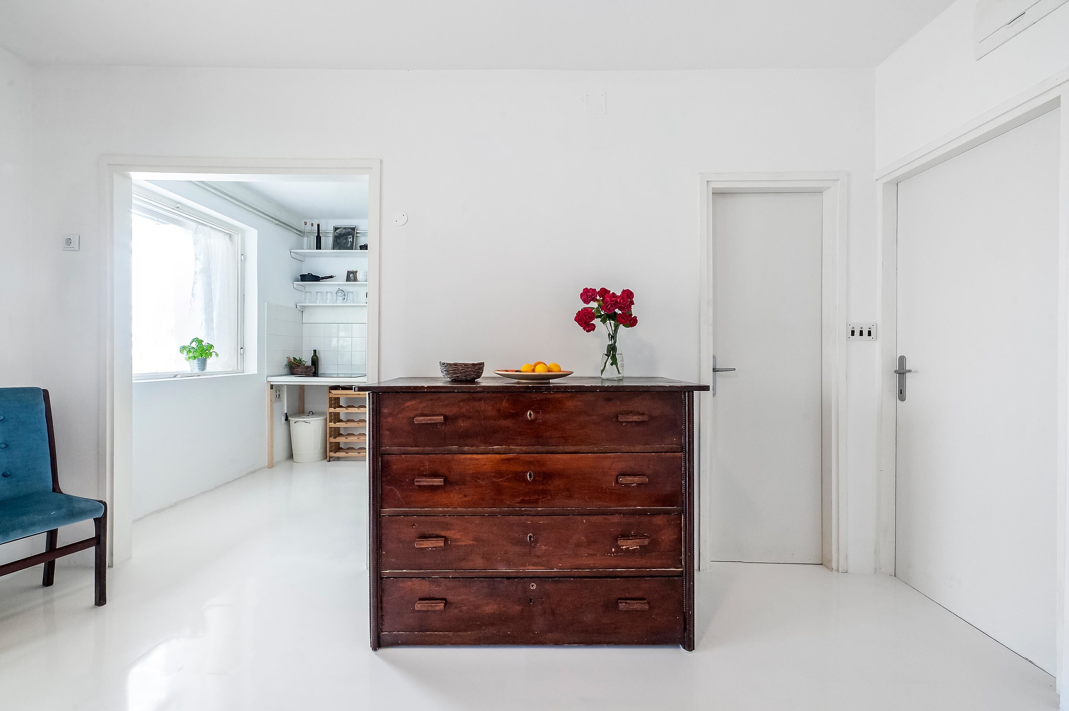 Una cocina moderna con paredes blancas, un aparador de madera, una silla azul y un jarrón con flores rojas en la encimera.