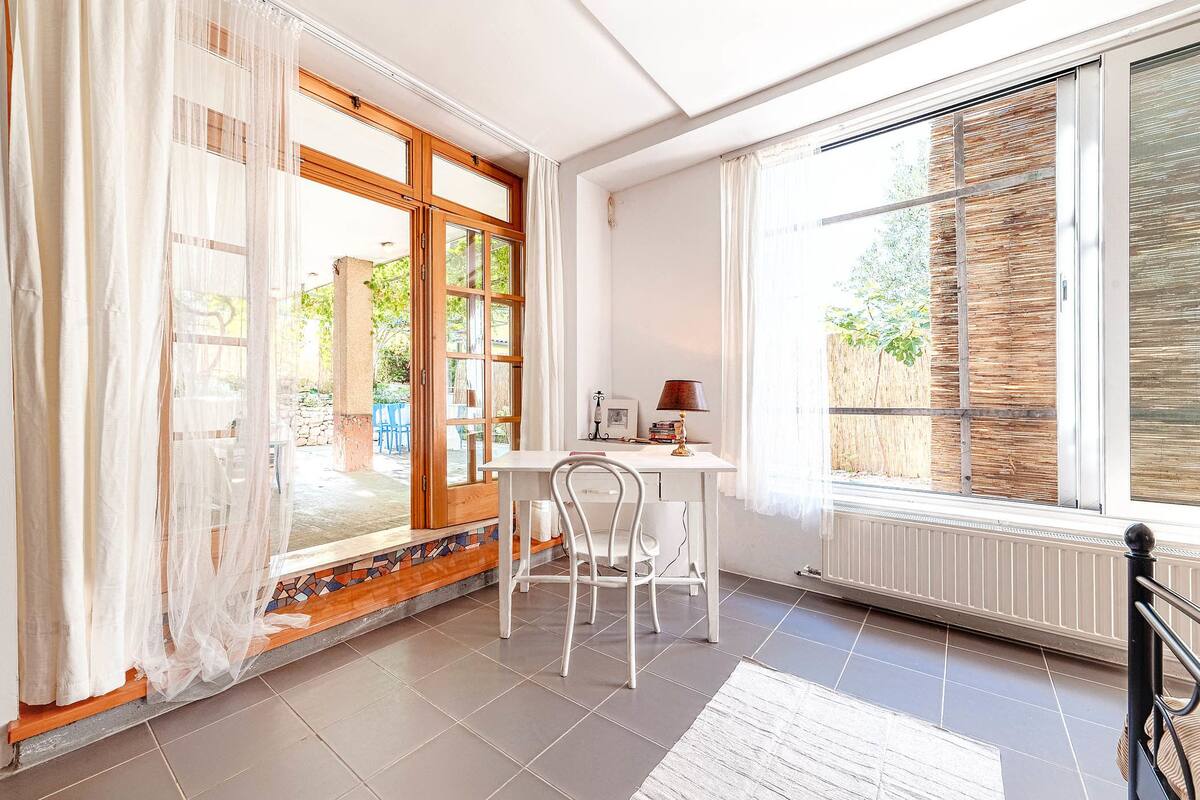 A bright room with a white desk and chair, large windows, and a view of greenery outside.