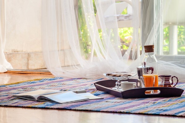 Una preparación para el desayuno con una bandeja, un libro, un vaso de jugo de naranja y una cafetera sobre una alfombra colorida cerca de una ventana con cortinas traslúcidas.