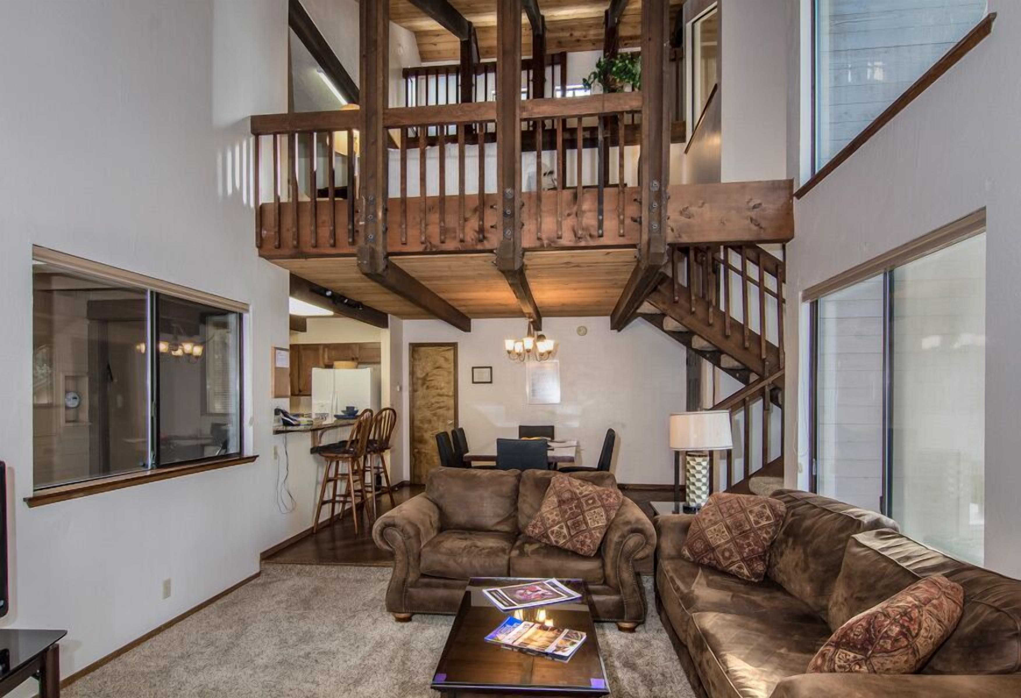 A spacious living room with a high wooden staircase, a large window, and a cozy seating area.