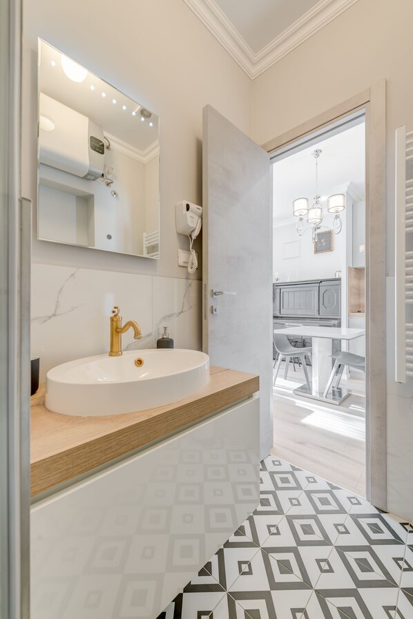 Un baño moderno con un lavamanos blanco, grifo dorado y un espejo grande. Hay un secador de pelo en la pared y una puerta que da a un comedor.