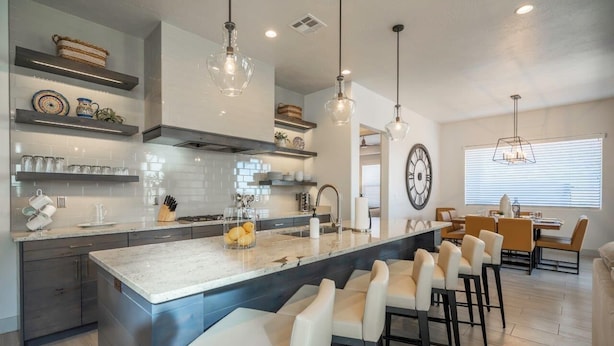 Cocina moderna con una isla grande, taburetes de bar, iluminación colgante y una zona de comedor con un reloj grande en la pared.