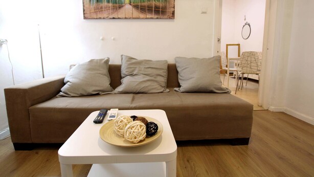 A living room with a brown sofa, a white coffee table with decorative items, and a painting on the wall. - KAV Apartments -Near Assuta Olei Gardom (Tel Aviv)